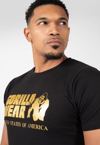 Fekete pamut póló arany szöveggel és egy gorilla grafikával. A dizájn tartalmazza a "GORILLA WEAR" feliratot és az "EGYESÜLT ÁLLAMOK" szöveget alatta.