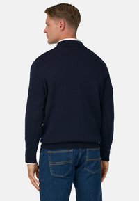 Jūras spēku adīts džemperis ar ribotiem piedurknēm un apakšmalu, apvienots ar zilām džinsām, kurām ir divas aizmugurējās kabatas un dzeltenu šuvju akcenti.