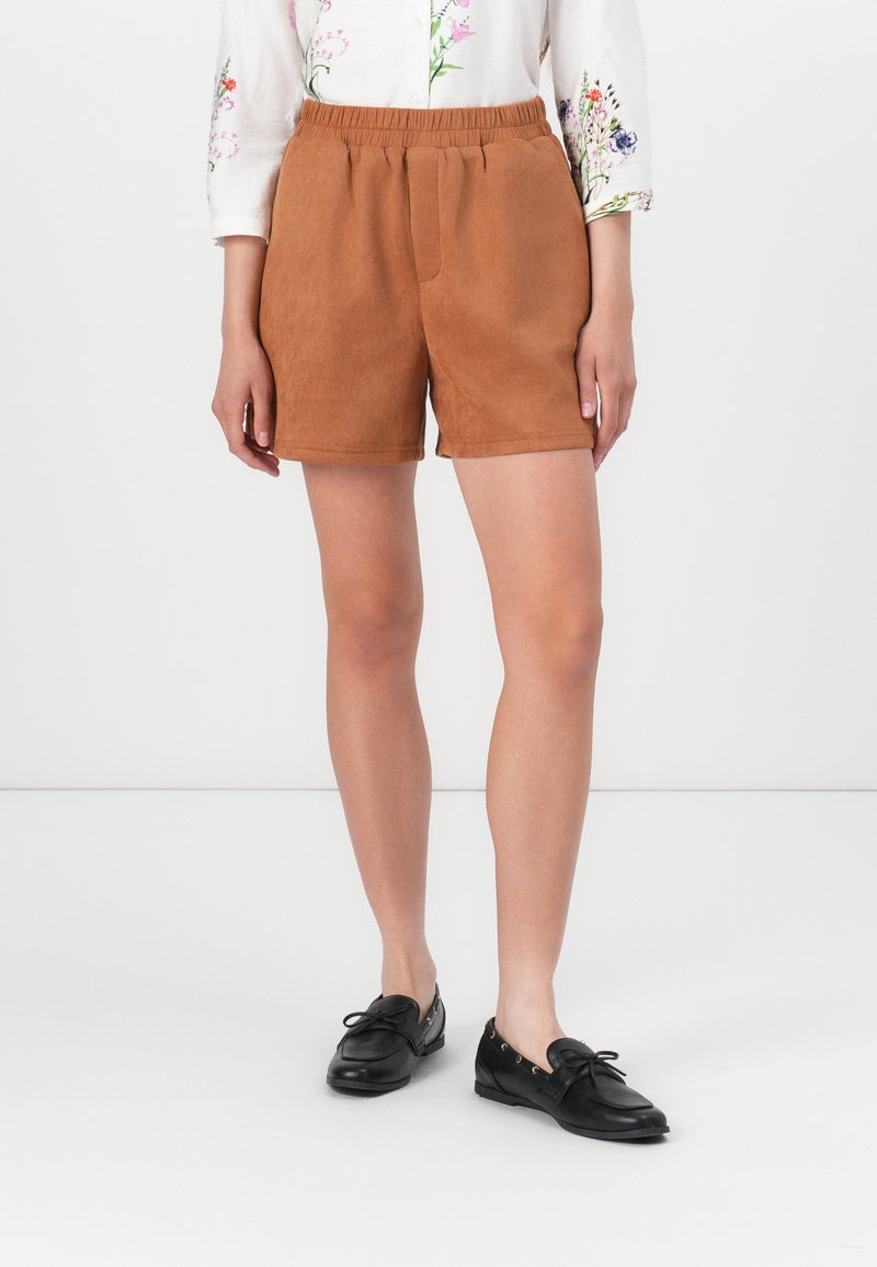 Bruine suède shorts met een elastische tailleband en zijzakken. Gecombineerd met zwarte instap schoenen, voorzien van een strikdetail en metalen accenten.