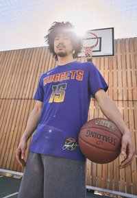 Violet krāsas basketbola krekls ar "NUGGETS 15" izpildījumā ar teksturētām zelta burtiem, apvienots ar pelēkiem šortiem un Spalding basketbolu. Koksnes fons.