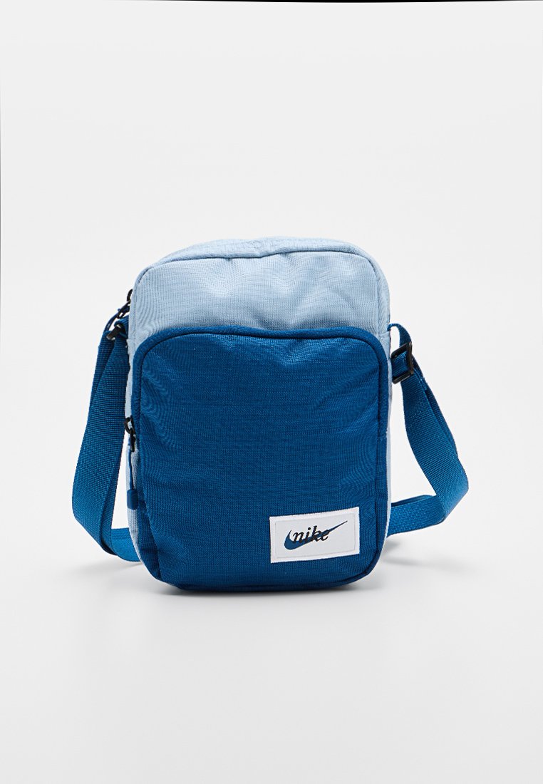 Bolso bandolera de tela azul con sección superior en un azul más claro, forma rectangular, correa ajustable y parche con logo blanco.
