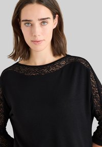 Pull noir avec un overlay en dentelle à motifs géométriques sur les épaules, coupe décontractée et texture douce. Col rond et manches longues.