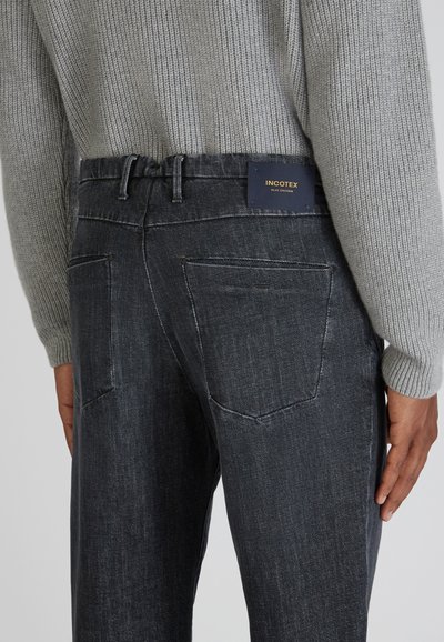 Pantaloni in denim grigio scuro con una consistenza liscia, dotati di due tasche posteriori e di un'etichetta con logo sulla vita. Maglione grigio a coste visibile.