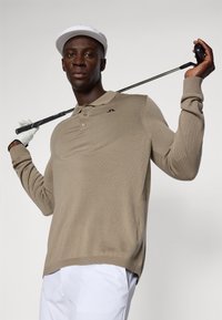 Beige kötött, hosszú ujjú pólóing, két gombos nyakkivágással és logó részlettel. A modell golfütővel pózol, fehér sapkát és kesztyűt visel.