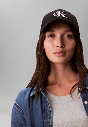 Joven mujer con gorra negra de Calvin Klein, camiseta gris con cuello de canalé y chaqueta vaquera azul sobre un fondo liso de color gris claro.