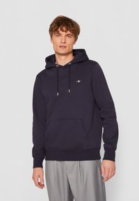 Marineblå hoodie med snore, frontlomme og lille logo detalje. Glat stof med ribbede manchetter og kant. Casual fit, praktisk design.