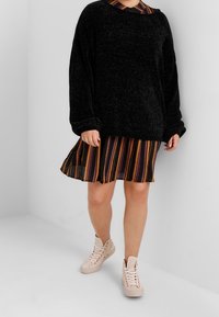 Maglione nero oversize in maglia indossato sopra un vestito a righe marrone, nero e arancione, abbinato a sneakers beige con lacci.