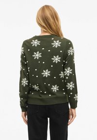 Woman with long blonde hair wearing a dark green sweater with white snowflake and dot patterns, paired with black pants, facing away.