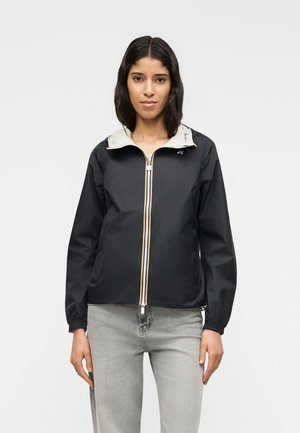 Femme portant une veste zippée noire avec une capuche gris clair et un jean gris clair, debout devant un fond blanc uni.