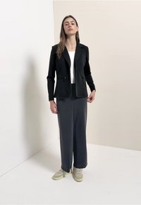 Blazer nero strutturato con tasche con zip, abbinato a una gonna lunga grigia e liscia e sneaker chiare. La modella posa su uno sfondo neutro.