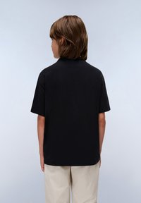 Svart bomullströja med korta ärmar och rund hals, visad bakifrån. Bärs med ljusa, strukturerade beige byxor.