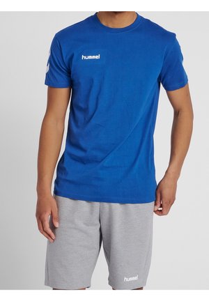 Homme portant un t-shirt bleu Hummel à manches courtes et un short gris clair Hummel, debout devant un fond uni.