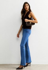 Top marrone senza maniche, jeans a zampa d'elefante blu e décolleté nere a punta. Gli accessori includono una piccola borsa marrone con un accento dorato. Materiali testurizzati visibili.