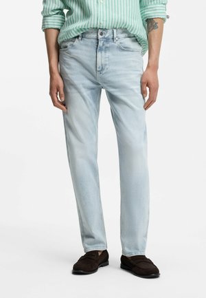 Mężczyzna w jasnoniebieskich dżinsach, brązowych mokasynach i zielono-białej koszuli w paski z podwiniętymi rękawami.