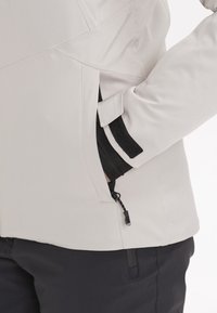 Veste gris clair avec poche latérale zippée et sangle Velcro sur la manche, portée sur des vêtements noirs, main dans la poche.