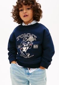 Νεαρό παιδί με σγουρά μαλλιά, φοράει μπλε σκούρο φούτερ με χαρακτήρα καρτούν σκύλου που παίζει μπάσκετ και ανοιχτό μπλε τζιν, με τα χέρια στις τσέπες.