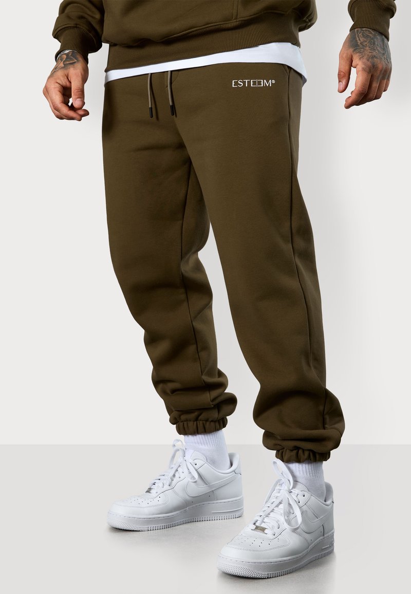 Olivgrüne Jogginghosen mit elastischen Bündchen und Kordelzug. Mit weißem Logo auf der Vorderseite. Kombiniert mit weißen Sneakers und sichtbaren weißen Socken.