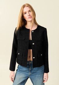 Schwarze strukturierte Jacke mit rundem Kragen, silbernen Knöpfen und zwei frontalen Taschen. Getragen über einem schwarzen Oberteil und hellblauen Jeans.