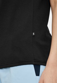 Camisa negra de manga corta hecha de una tela suave, con una etiqueta en el dobladillo lateral que presenta un logotipo circular. Textura suave con un ajuste regular.