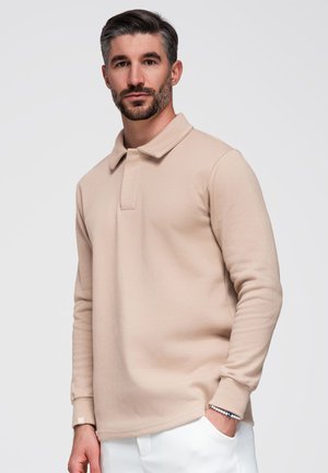 Man met kort donker haar en baard draagt een beige overhemd met lange mouwen en kraag en een witte broek, staand tegen een effen lichte achtergrond.