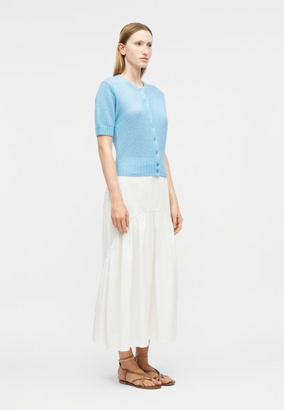 Femme debout de profil portant un cardigan boutonné à manches courtes bleu clair, une jupe blanche jusqu'aux chevilles et des sandales marron à brides.