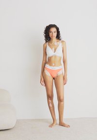 Witte bralette en oranje bikini bottoms met logo Tailleband. Glad materiaal, eenvoudig ontwerp en driehoekige cupvorm. Model staat op een tapijt.