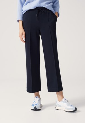 Person iført løse sorte cropped bukser, lyseblå sweater og hvide sneakers med blå detaljer, står på en lys baggrund.