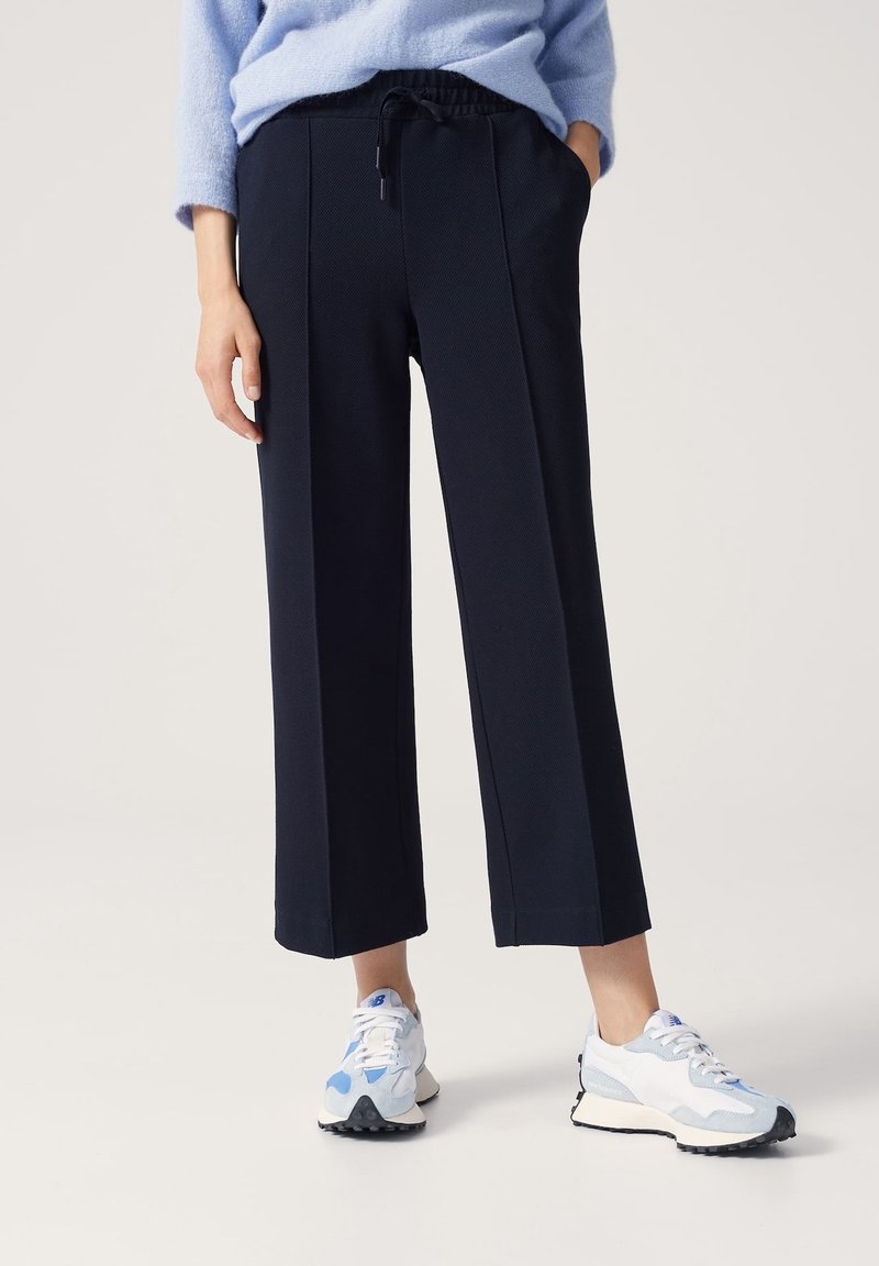 Personne portant un pantalon noir ample et raccourci, un pull bleu clair et des baskets blanches avec des accents bleus, debout sur un fond clair.
