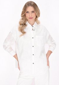 Weißes Button-Up-Hemd mit floraler Textur, schwarzen Knöpfen und einer Brusttasche. Langärmlig, die Ärmel hochgerollt für einen lässigen Sitz vor einem hellen Hintergrund.