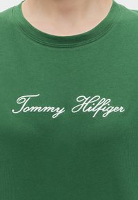 Gros plan d'une chemise verte avec un texte brodé en cursive blanche indiquant "Tommy Hilfiger" sur la poitrine sous un col rond.