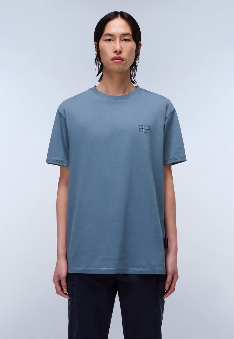 Camiseta de algodón azul claro con cuello redondo, mangas cortas y un pequeño logotipo en relieve en el pecho. Tejido texturizado, corte relajado.