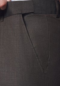 Brązowa, teksturowana tkanina dopasowanych spodni, widoczna kieszeń boczna i pas. Materiał wydaje się wytrzymały o gładkim wykończeniu.