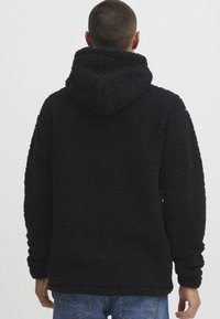 Svart fleece hettegenser med et teksturert mønster og en stor hette. Har en avslappet passform og lange ermer. Bært over blå jeans.