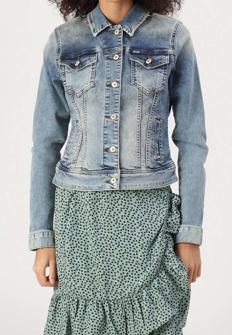 Chaqueta de mezclilla de color azul claro con un diseño ajustado, dos bolsillos en el pecho, herrajes plateados y una textura ligeramente desgastada. Combinada con una falda verde de estampado.