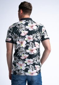 Camisa polo de manga corta negra con grandes patrones florales tropicales en blanco y rosa, con un tejido texturizado y un cuello negro sólido.