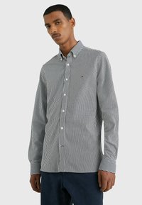 Camisa de manga larga a rayas en blanco y negro con cuello abotonado, botones blancos y pequeño detalle de logo en el pecho. Tejido suave y de sarga.