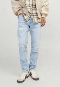 Lichtblauwe denim jeans met versleten details, rechte snit, gedragen met een beige bomberjack en een geruit shirt, gecombineerd met witte sneakers.