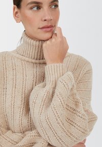 Beige gestrickter Pullover mit einem hohen Rippkragen, weiten Ärmeln und strukturierten Streifen. Besitzt subtile goldene Pailletten für zusätzliche Details.
