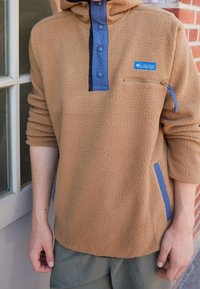 Pullover in fleece color beige con dettagli blu. Presenta una tasca con zip, una patta con bottoni e un tessuto testurizzato. Vestibilità comoda e maniche lunghe.