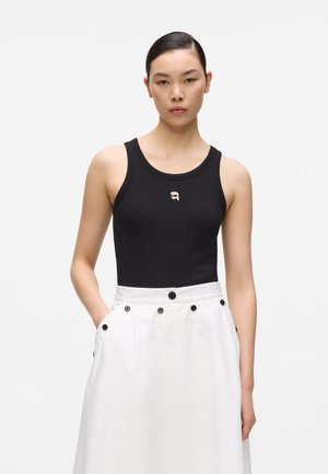 Jeune femme portant un haut noir sans manches ajusté avec un petit visage brodé et une jupe blanche taille haute avec des boutons noirs.