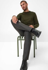 Grijze, textuurstof broek met een slim fit, gecombineerd met een donkergroen, langemouwenshirt. Zwarte veterschoenen. Model zit op een groene stoel.