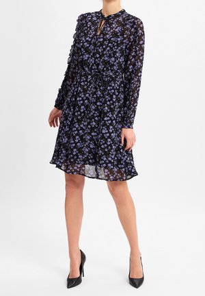 Frau trägt ein schwarzes, durchsichtiges Kleid mit lila Blumen, langen Ärmeln, gebundener Taille und schwarzen High Heels, steht vor weißem Hintergrund.