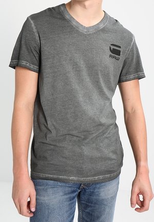 Personne portant un t-shirt gris foncé délavé à col en V avec un logo G RAW sur la poitrine et un jean bleu, debout devant un fond uni.