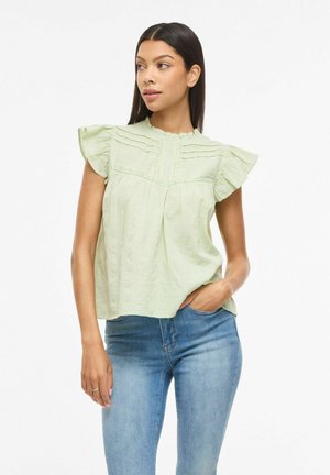 Žena v svetlozelenom blúzke s krátkymi volánikovými rukávmi a čipkovými detailmi, modrých džínsoch, stojaca s jednou rukou v vrecku.