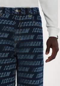 Denim jeans featuring a dark blue color with an all-over repeating "ICECREAM" print in varying shades. Two visible front pockets and yellow stitching.
