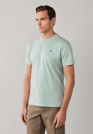Man draagt lichtgroen T-shirt met korte mouwen en klein donkerblauw logo op de borst en beige broek, staand tegen een effen lichtgrijze achtergrond.