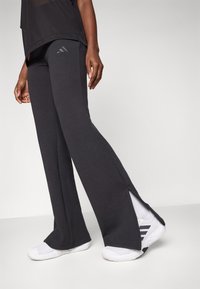 Σκούρα γκρι αθλητικά παντελόνια με άνοιγμα στον πλάι, κατασκευασμένα από μαλακό ύφασμα. Διαθέτουν λογότυπο της Adidas στη ζώνη και είναι συνδυασμένα με λευκά αθλητικά παπούτσια.