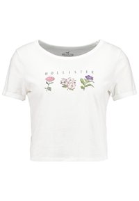 White cropped t-shirt made of cotton featuring four floral illustrations in pink and purple with "Hollister" text underneath. Short sleeves.