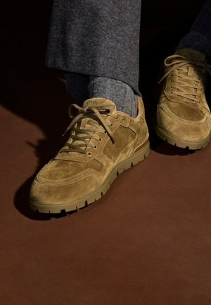 Tan suede sneakers met dikke rubberen zolen, gedragen met grijze sokken en donkergrijze broeken tegen een donkere achtergrond.