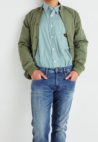 Ljus olivgrön, vadderad bomberjacka över en blå och vit randig skjorta, kombinerad med urblekta blå jeans. Händerna i fickorna.